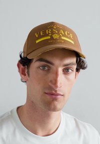 Căciulă maro Versace cu text și logo brodat în galben, brădiță curbă și curea ajustabilă la spate, purtată de o persoană cu o cămașă albă.