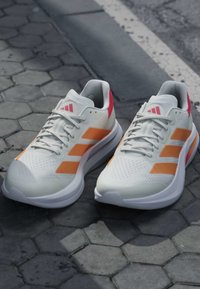 Paire de chaussures de sport blanches avec des bandes orange et des accents roses sur un pavé hexagonal près d'un trottoir.