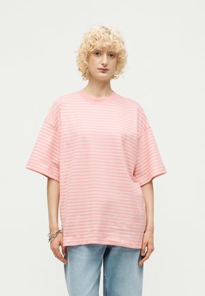 Osoba o kręconych blond włosach, ubrana w luźną różowo-białą koszulę w paski z krótkim rękawem oraz jasnoniebieskie dżinsy na jednolitym tle.