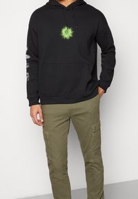 Sudadera con capucha negra de algodón, con un diseño de corazón verde brillante en el pecho y gráficos en las mangas; combinada con pantalones cargo color oliva.