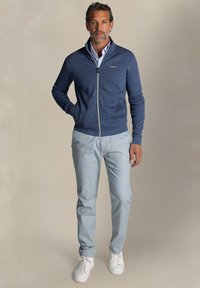Marine blauwe zip-up trui met een hoge kraag, een lichtblauw shirt eronder, lichtblauwe broek en witte sneakers. Details omvatten een logo op de trui.