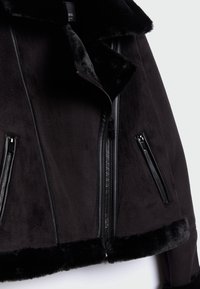 Veste en fausse fourrure noire avec une texture douce, dotée d'un large col, d'accents en cuir et de plusieurs poches zippées à l'avant.