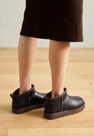 Bottines en cuir marron avec doublure en peluche, languettes de tirage et semelle texturée, associées à une jupe marron côtelée sur un sol en bois.