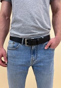 Ceinture en cuir noir avec une boucle en métal argenté, portée sur un jean bleu clair, présentant une texture lisse et un design classique. Un t-shirt gris est visible au-dessus.