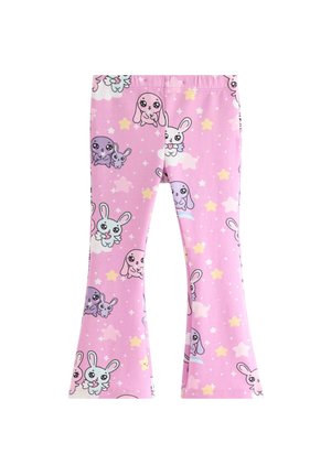 Ung pige med rødt hår iført pink pyjamasæt med tegneserie-kaniner og stjernemønstre, smilende og stående foran en ensfarvet baggrund.