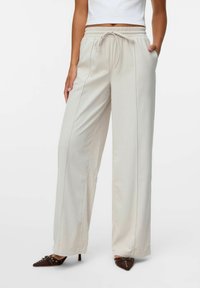 Pantalon ample de couleur crème avec un tissu texturé, taille élastique et poches latérales. Associé à des chaussures à bout pointu marron.