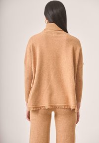 Suéter de punto beige, de gran tamaño, con cuello alto, que presenta un dobladillo deshilachado y detalles acanalados. Textura suave, diseñado para la comodidad.