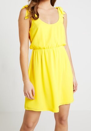 Kobieta ubrana w żółtą sukienkę bez rękawów z wiązanymi ramiączkami i elastycznym taliowaniem, stojąca na jednolitym białym tle.