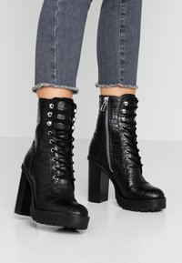 Czarne botki na obcasie sięgające kostki z teksturą krokodyla, z przodem na sznurowanie, zamkiem błyskawicznym z boku oraz grubą platformą. Dżinsowe spodnie z postrzępionymi mankietami.