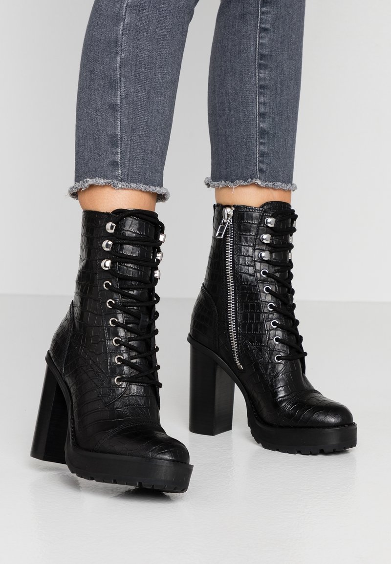 Czarne botki na obcasie sięgające kostki z teksturą krokodyla, z przodem na sznurowanie, zamkiem błyskawicznym z boku oraz grubą platformą. Dżinsowe spodnie z postrzępionymi mankietami.