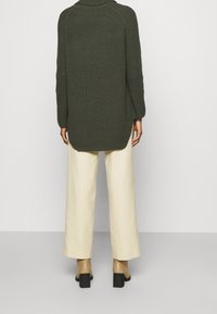 Tmavě zelený pletený svetr s vysokým límcem a dlouhými rukávy, kombinovaný s krémovými širokými kalhotami a světle hnědými botami na širokém podpatku.