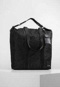 Zwarte nylon tote bag met dubbele handvatten, verstelbare schouderband, verticale naden en een merkembleem aan de voorkant.