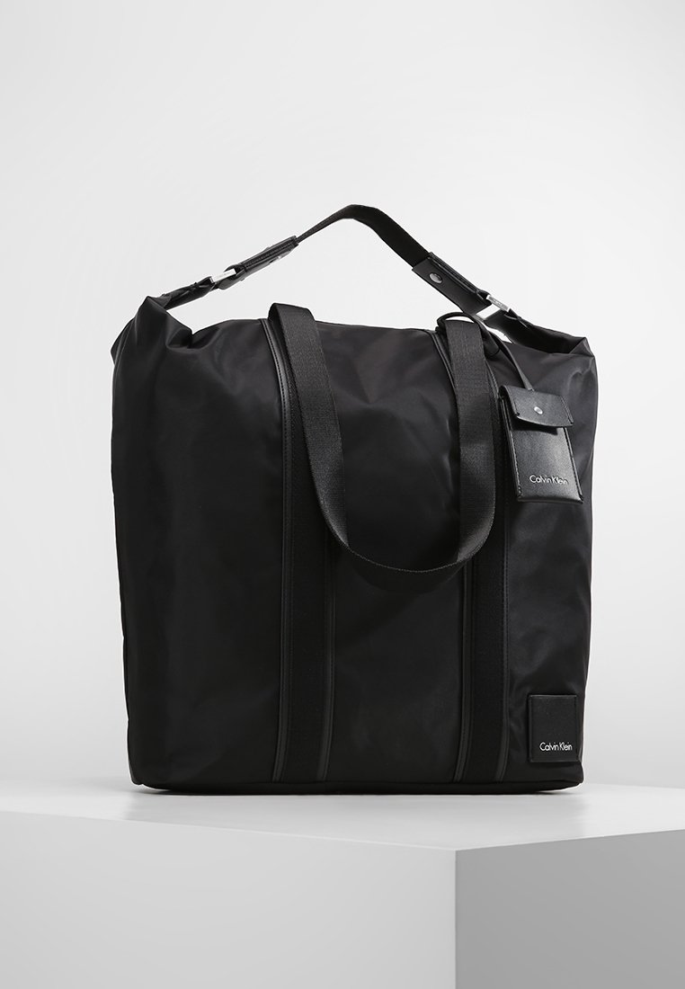 Zwarte nylon tote bag met dubbele handvatten, verstelbare schouderband, verticale naden en een merkembleem aan de voorkant.