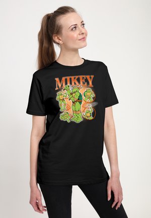Jeune femme portant un t-shirt noir avec plusieurs personnages de dessin animé des Tortues Ninja et le mot « MIKEY » en orange au-dessus.