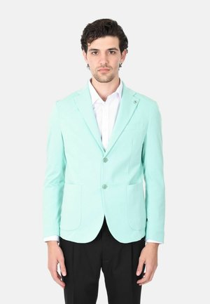 Uomo in piedi con volto rivolto in avanti, indossa un blazer verde menta sopra una camicia bianca e pantaloni neri su uno sfondo chiaro uniforme.
