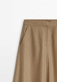 Pantalon en laine marron camel taille haute avec poches latérales et fermeture éclair dissimulée à l'avant, présenté sur fond blanc.