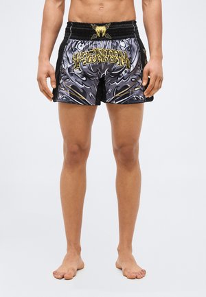 Άνδρας με μαύρο και γκρι σορτς Muay Thai με χρυσό κείμενο "Venum" και σχέδιο, στέκεται ξυπόλυτος σε απλό φωτεινό φόντο.