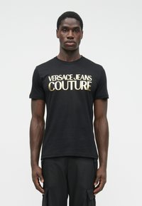 Stāvošs vīrietis, tērpies melnā T-kreklā ar uzrakstu "VERSACE JEANS COUTURE" zeltainiem burtiem un melnās kravas biksēs pret vienkāršu fonu.