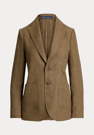 Femme debout, les mains dans les poches, portant un costume à carreaux marron, une chemise beige, une ceinture noire et des chaussures marron sur un fond neutre.