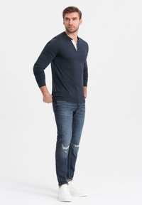 Marineblaues Langarm-Henley-Shirt mit dreiknöpfiger Öffnung, kombiniert mit abgenutzten dunkelblauen Jeans und weißen Sneakers, auf einem Modell vor einem schlichten Hintergrund.