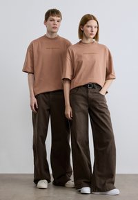 Δύο άτομα φοράνε υπερμεγέθη, σκουροκόκκινα μπλουζάκια με το κείμενο «BROKE SINCE MILANO», συνδυασμένα με φαρδιά, σκούρα καφέ παντελόνια και λευκά παπούτσια.