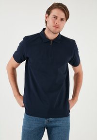 Námořnická modrá polo košile s krátkým rukávem, s límcem na zip a hladkou texturou. Nesená s světle modrými džíny.