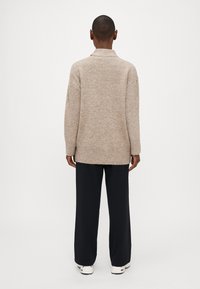 Maglione beige a maglia con collo alto, polsini e orlo a coste, abbinato a pantaloni neri a gamba larga e sneaker bianche.