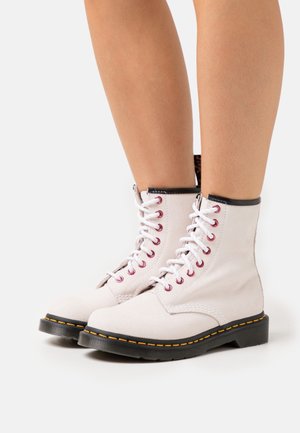 Bottes de combat lacées blanches en cuir texturé, avec des œillets rouges, une bordure noire et des coutures jaunes le long de la semelle.