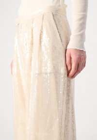 Beige, met pailletten bezette stoffen broek met een geplooid ontwerp en een tailleband. De pailletten creëren een reflecterende textuur, wat de visuele details versterkt.