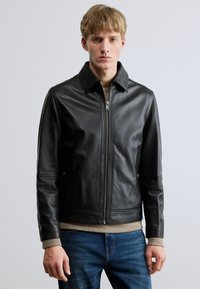 Fekete bőr dzseki cipzáros elülső résszel, lapos gallérral és bordázott mandzsettákkal. Simább textúrájú és testhezálló kialakítású, bézs pulóverrel párosítva.