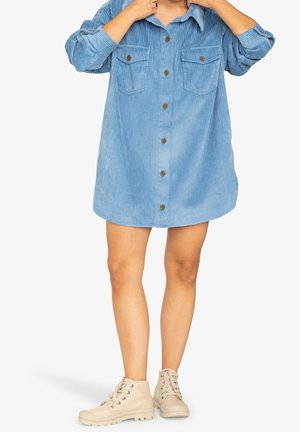 Kvinde iført oversized blå kordfløjlskjole med brune knapper og beige høje sneakers på hvid baggrund.