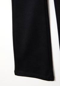 Primer plano de la parte inferior de la pierna de unos pantalones de tela negra con costuras visibles, contra un fondo blanco.