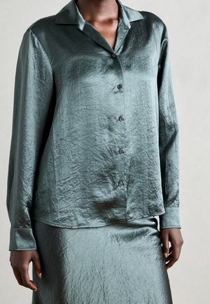 Vrouw draagt een gerimpelde teal lange mouwen blouse met knopen en bijpassende rok, staand tegen een effen lichte achtergrond.