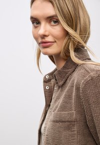 Camisa chaqueta de pana marrón con cuello, cierre de botones, bolsillo en el lado izquierdo y tela texturizada de canalé.