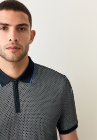 Sötét mintás galléros póló cipzárral az elején. Texturált mintázattal, rövid ujjakkal és kontrasztos kék szegéllyel a galléron.
