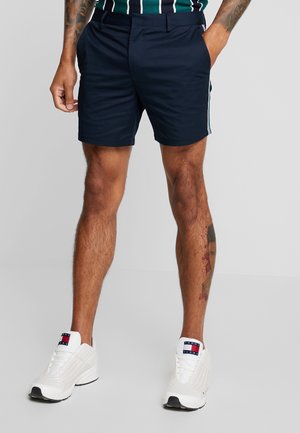 Marineblaue Shorts mit Seitenstreifen aus strapazierfähigem Stoff. Mit flacher Vorderseite, Gürtelschlaufen und Taschen. Kombiniert mit weißen Sportschuhen.