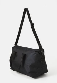 Geantă duffle neagră, realizată dintr-un textil rezistent. Dispune de două mâneri superioare, un curea de umăr ajustabilă și o închidere cu fermoar. Textură netedă.