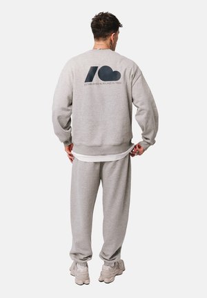 Mężczyzna w jasnoszarej bluzie i dresach, stojący tyłem do kamery, czarne logo i tekst na plecach bluzy, białe sportowe buty.