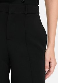 Pantaloni neri su misura in tessuto morbido, con tasche laterali e vita piatta, mostrati con una mano appoggiata all'anca.