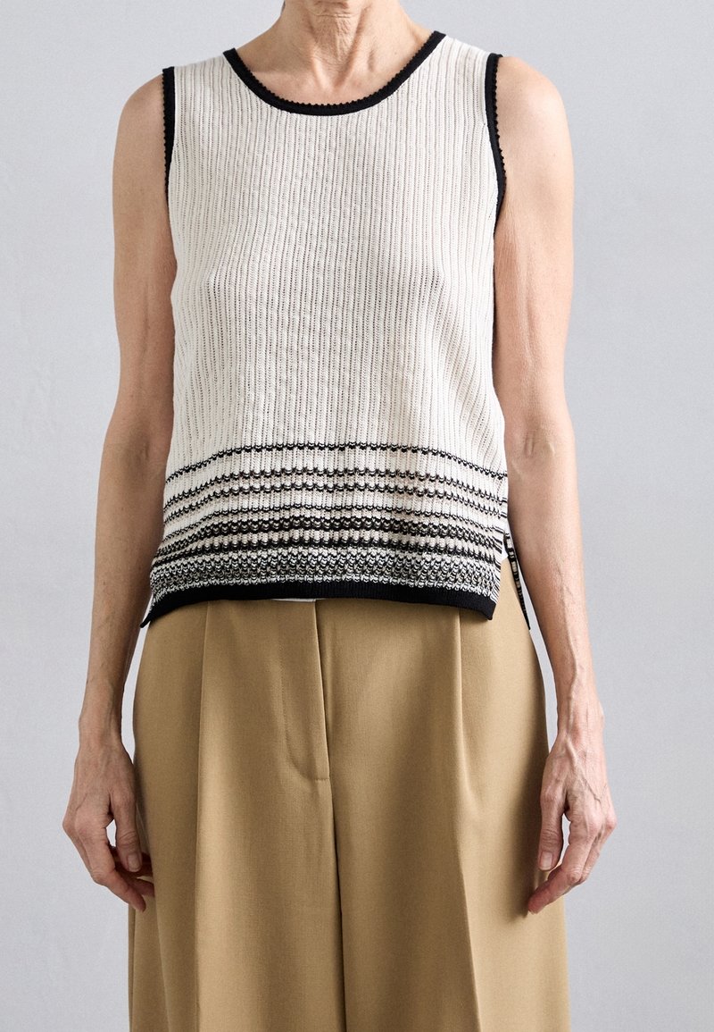 Haut sans manches en maille blanche avec bordure noire et rayures horizontales noires près de l'ourlet, porté avec un pantalon beige taille haute.