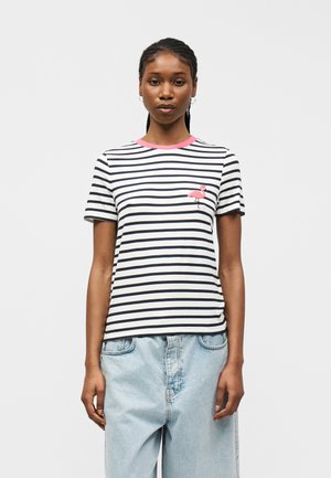 Γυναίκα που στέκεται με πρόσωπο προς τα εμπρός, φοράει λευκό και μαύρο ριγέ μπλουζάκι με ροζ πατς φλαμίνγκο και ανοιχτό μπλε τζιν παντελόνι.