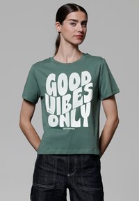 Groene katoenen t-shirt met korte mouwen, voorzien van grote witte tekst "GOOD VIBES ONLY" in een vet, speels lettertype.