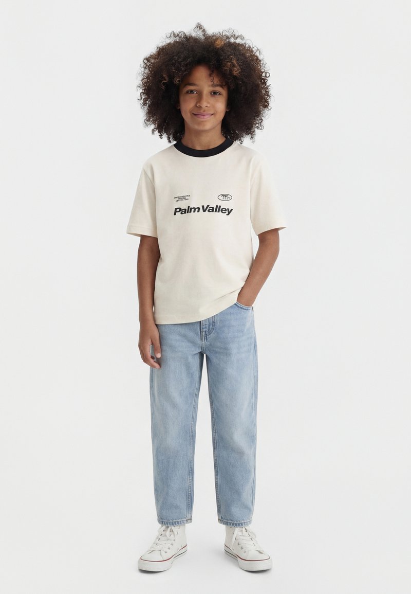 Child with curly hair wearing a cream "Palm Valley" t-shirt, light blue jeans, and white sneakers, standing with one hand in pocket.