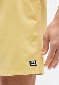 Κοντινό πλάνο ενός χεριού δίπλα σε κίτρινα σορτς με ένα μικρό μαύρο και άσπρο λογότυπο κυμάτων κοντά στο τελείωμα.