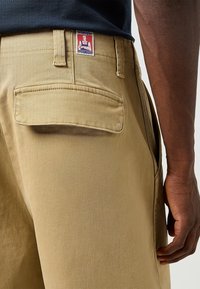 Pantalones Wrangler de color caqui con una textura suave, que cuentan con un bolsillo trasero y una etiqueta de marca. Mano y brazo visibles en el encuadre.