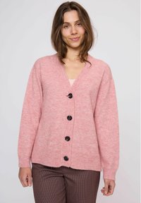 Roze wollen cardigan met een v-hals, vijf zwarte knopen, geribbelde zoom en manchetten, en een zachte textuur. Gekleed over een licht topje en een patroondroge broek.