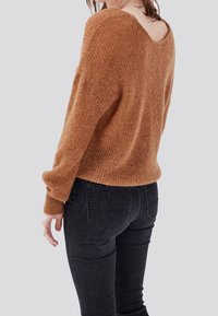 Femme portant un pull en peluche marron avec un décolleté en V dans le dos et un jean noir ajusté, debout devant un fond clair uni.