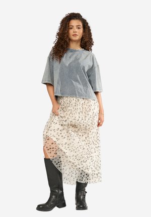 Mladá žena s kučeravými vlasmi, oblečená v sivej nadrozmernej tričku, bielej plisovanej sukni s čiernymi škvrnami a čiernych čižmách po kolená.