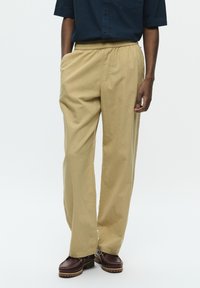 Pantaloni bej, cu picioare largi, având un brâu elastic și buzunare laterale, asortați cu pantofi maro închis cu o talpă robustă.