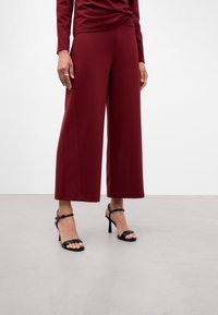 Pantaloni larghi bordeaux realizzati in tessuto morbido, con una texture liscia e una vestibilità rilassata. Abbinati a sandali neri con cinturini e tacco alto.